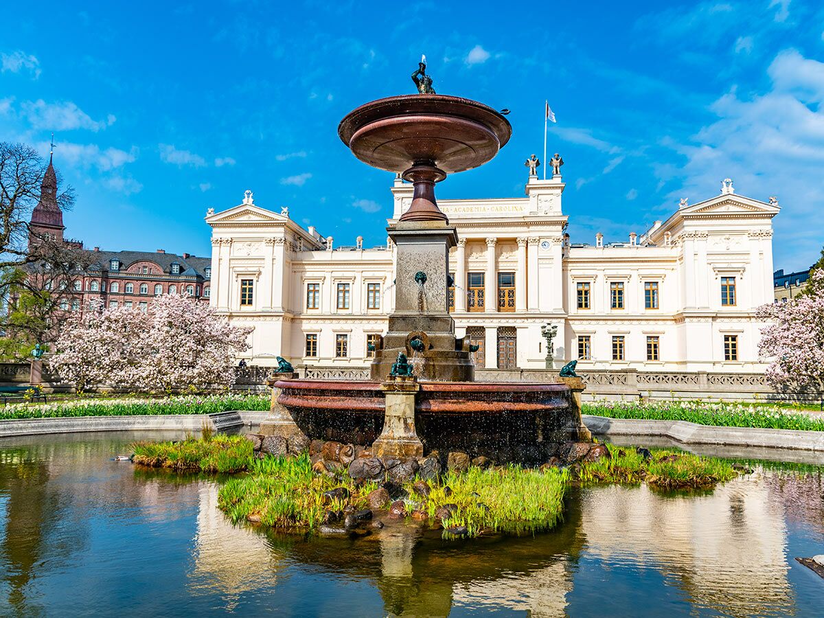Barockbrunnen vor dem Hauptgebäude der Universität Lund in Schweden im Frühling, mit blühenden Bäumen und blauem Himmel.