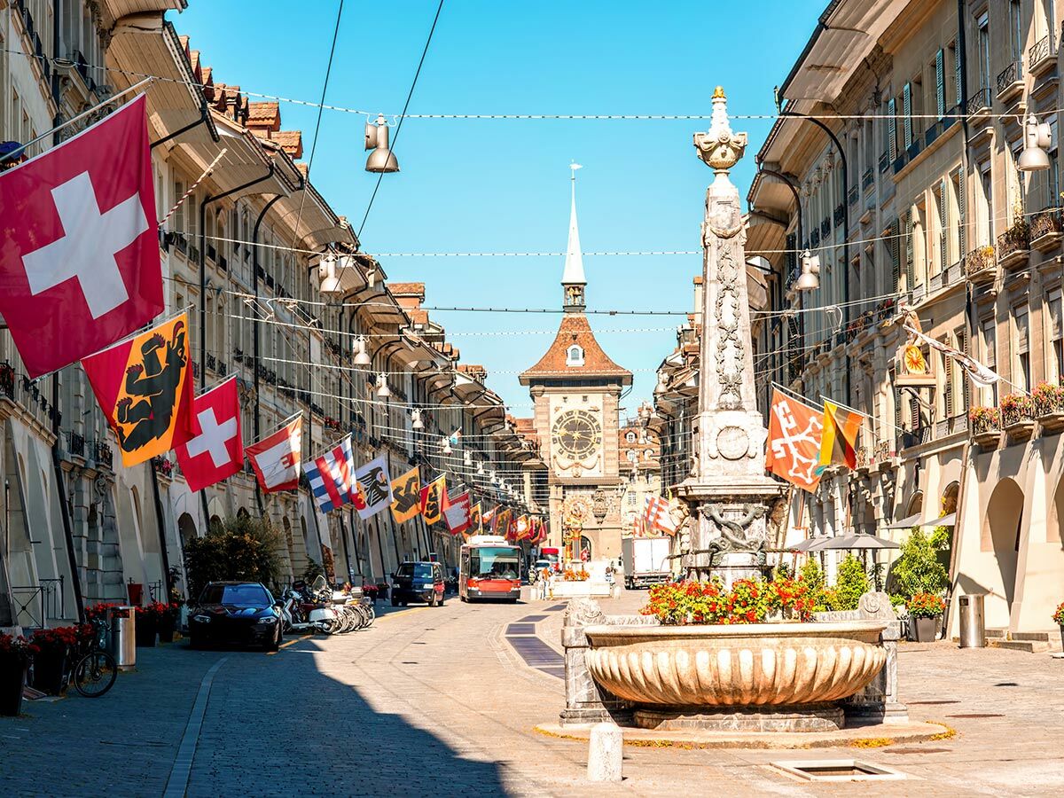 Blick auf die historische Zytglogge in der Berner Altstadt, geschmückt mit Schweizer und Kantonsfahnen an einem sonnigen Tag.