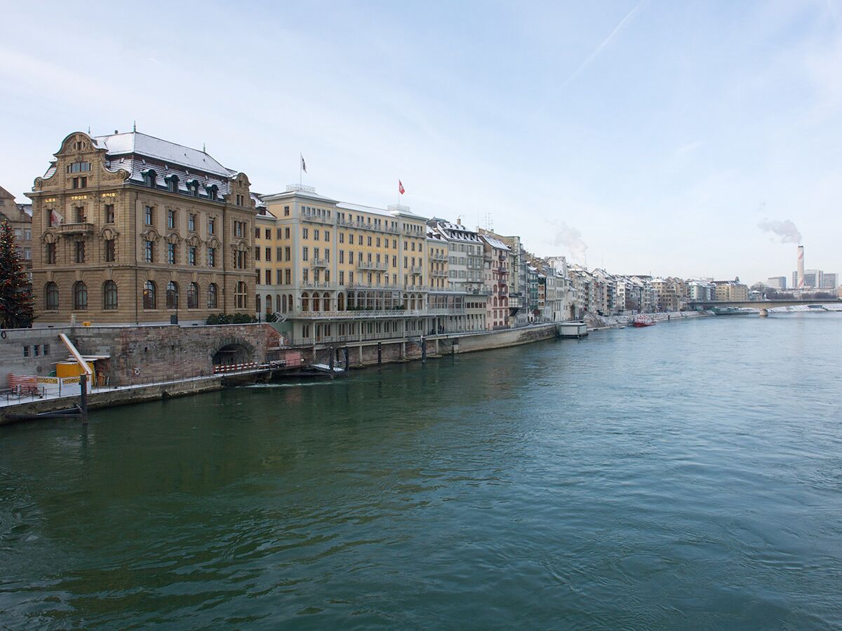 Blick auf die Uferpromenade von Basel am Rhein im Winter, mit schneebedeckten Dächern, historischen Gebäuden und Weihnachtbaum.