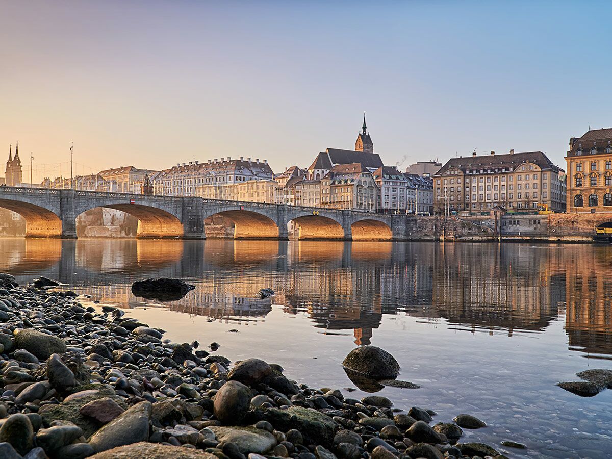 Blick auf Basel mit der Mittleren Brücke über den Rhein, historische Gebäude spiegeln sich im Wasser bei Sonnenaufgang.