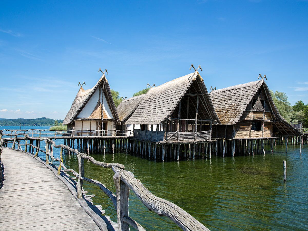 Historische Pfahlbauten am Bodensee, umgeben von klarem Wasser und einem Holzsteg, unter blauem Himmel.