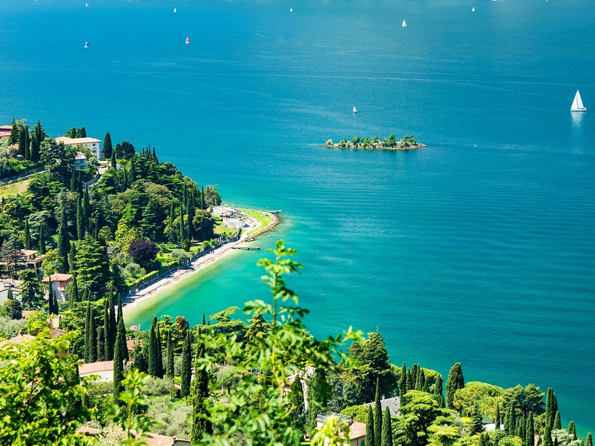 Panoramablick auf den Gardasee in Italien mit grünen Ufern, Villen, einer kleinen Insel und Segelbooten auf blauem Wasser.