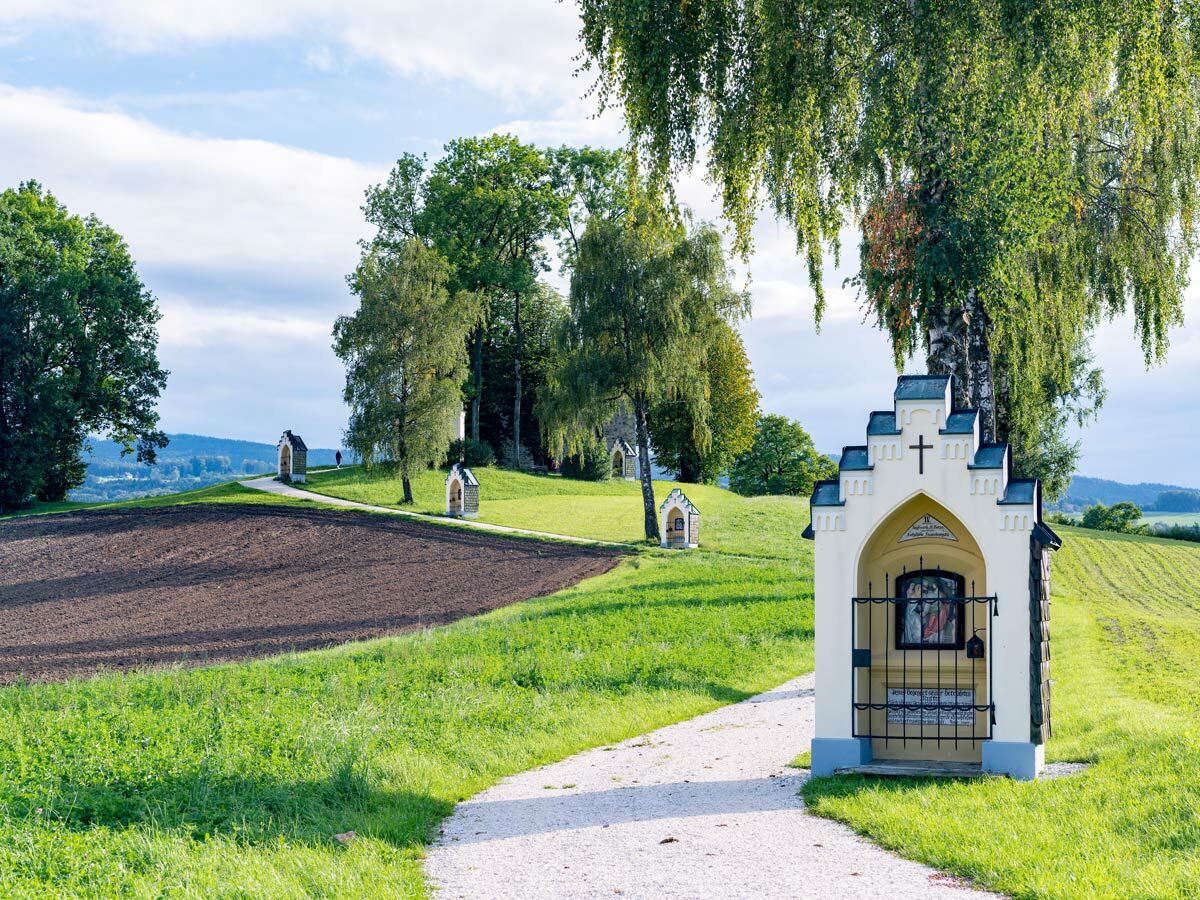 Reihe von historischen Bildstöcken mit religiösen Darstellungen säumt einen Schotterweg durch hügelige, grüne Landschaft unter blauem Himmel.