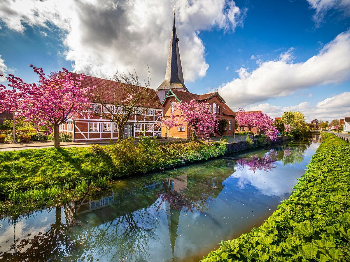 Idyllisches Dorf mit historischen Häusern, Kirchturm und rosa Kirschblütenbäumen entlang eines Kanals, im Wasser spiegelnd.