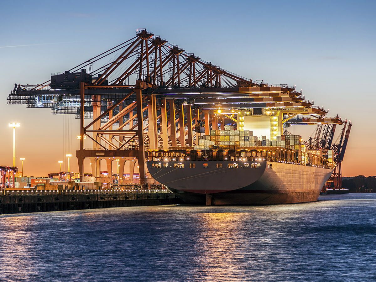 Großes Containerschiff liegt bei Dämmerung im beleuchteten Hafen. Zahlreiche Containerkräne laden Frachtgut auf das Schiff.