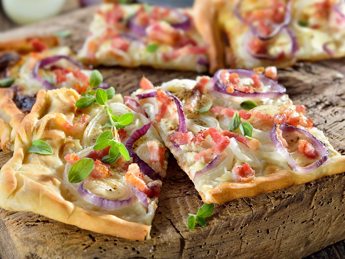 Mehrere Stücke Flammkuchen mit Zwiebeln, Speck und frischen Kräutern auf rustikalem Holzbrett. Ansicht von oben.