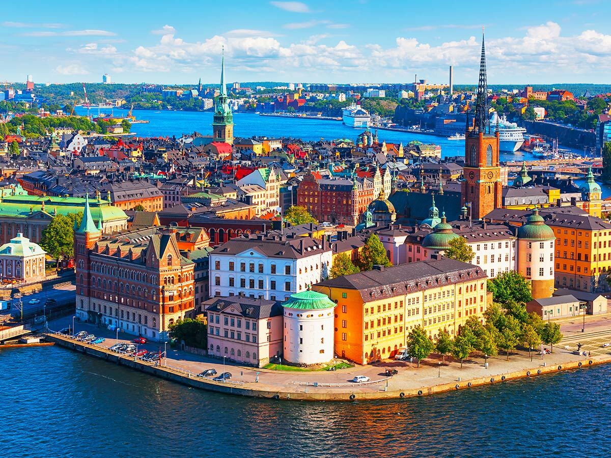 Luftaufnahme von Stockholms historischer Altstadt Gamla Stan am Wasser, mit bunten Gebäuden, Kirchtürmen und Kreuzfahrtschiffen bei Sonnenschein.