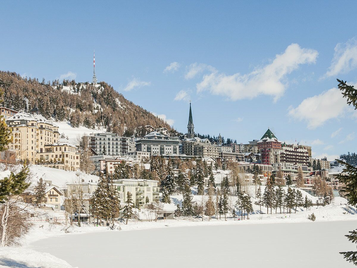 Panoramablick auf St. Moritz im Winter mit schneebedeckten Gebäuden, gefrorenem See und Bergen unter blauem Himmel.