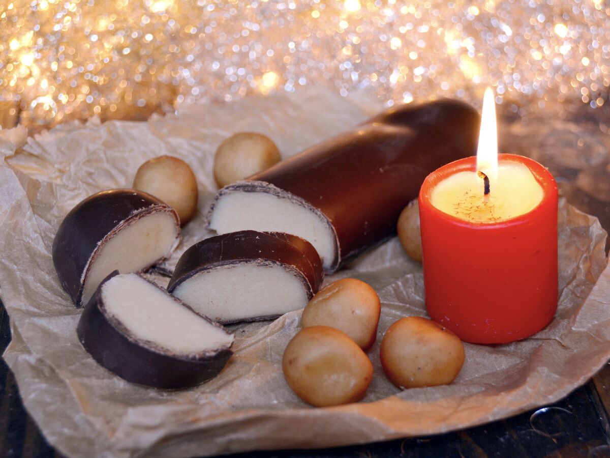 Geschnittene Marzipan-Schokoladenstangen und Marzipankartoffeln auf Packpapier, daneben eine brennende rote Kerze vor goldenem Bokeh-Hintergrund.