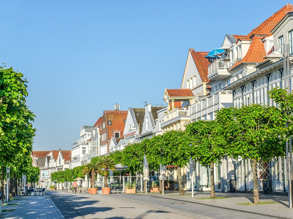 Sonnenbeschienene Promenade mit historischen Seebadgebäuden und begrünten Bäumen unter klarem blauen Himmel.