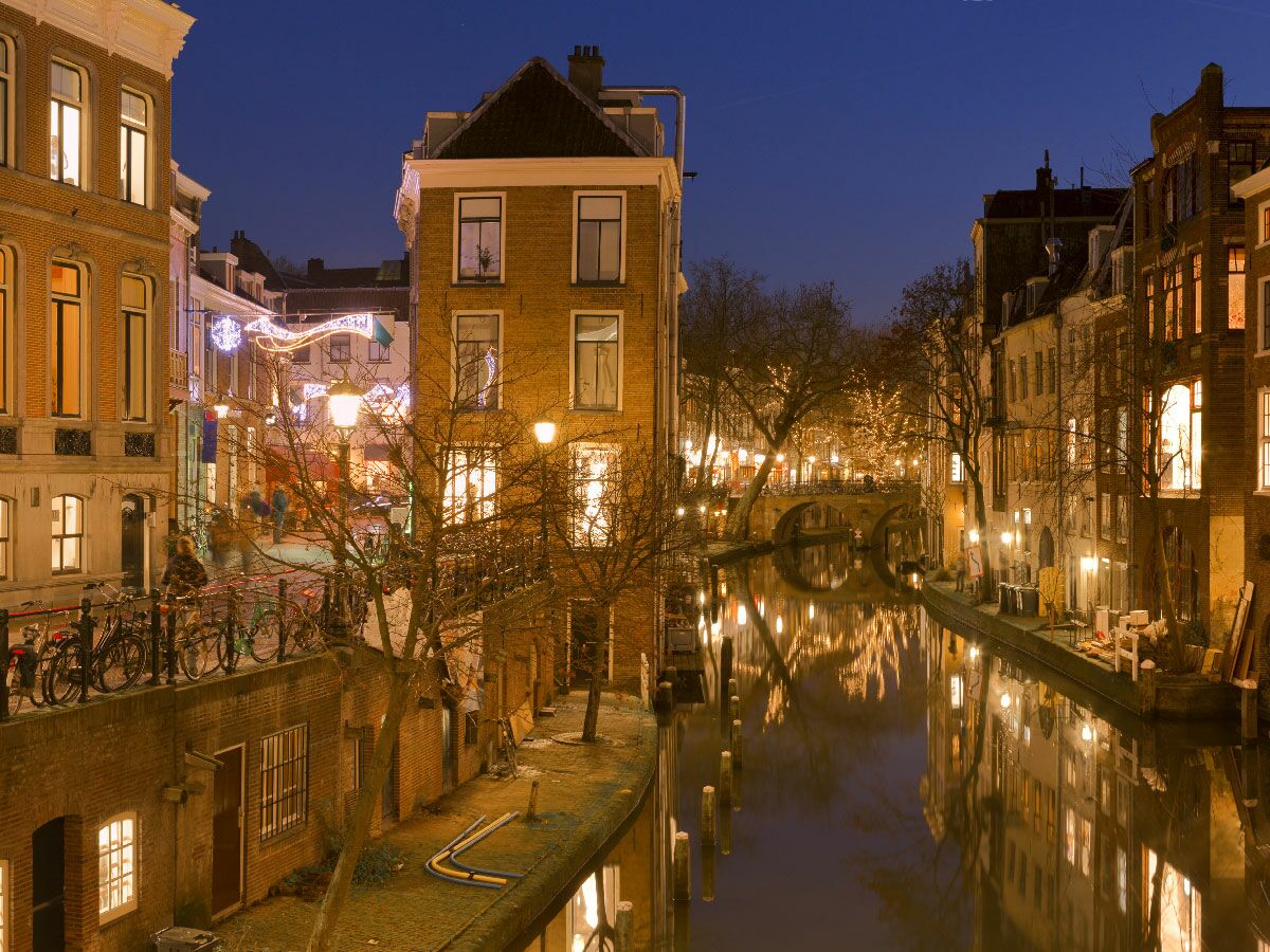 Nachtaufnahme des Oudegracht Kanals in Utrecht mit beleuchteten Gebäuden, Brücken und weihnachtlichen Lichtern, deren Reflexionen im Wasser.