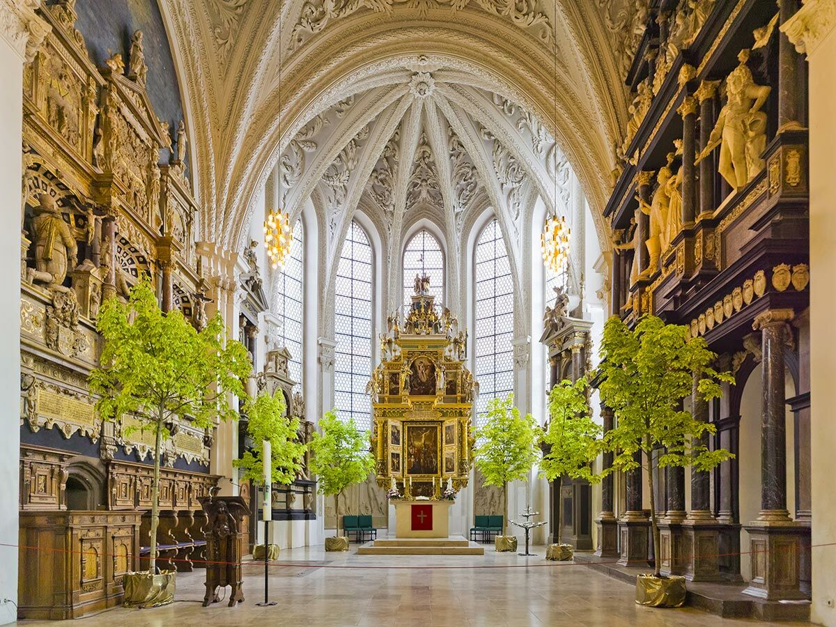 Prächtiger Kircheninnenraum mit hohem Altar, verzierten Gewölbedecken, großen Fenstern und dekorativen grünen Bäumen auf dem Boden.