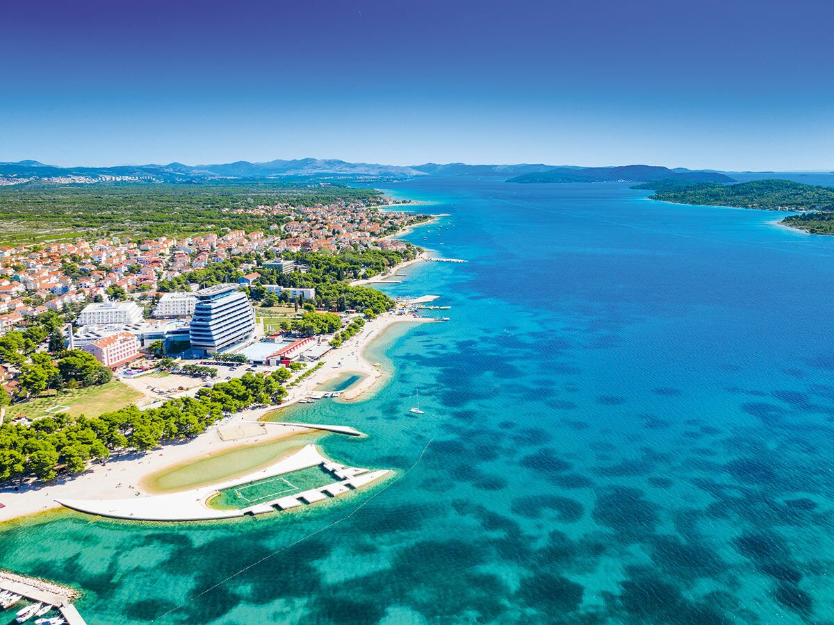 Luftaufnahme einer kroatischen Küstenstadt mit modernen Hotels, Sandstrand und türkisblauem Meer. Inseln im Hintergrund.