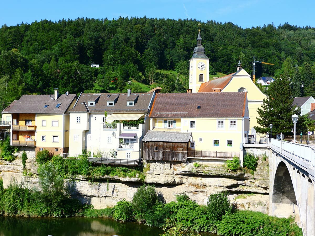 Blick auf eine historische Stadt am Flussufer mit bunten Häusern, einer Kirche mit Glockenturm und einer Steinbrücke.