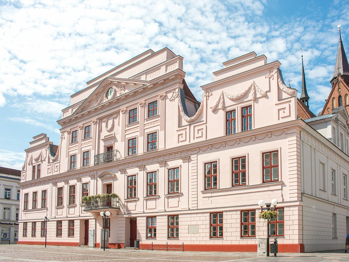 Großes historisches Rathaus mit rosa Fassade, dekorativen Giebeln und Uhr unter blauem Himmel in deutscher Stadt.