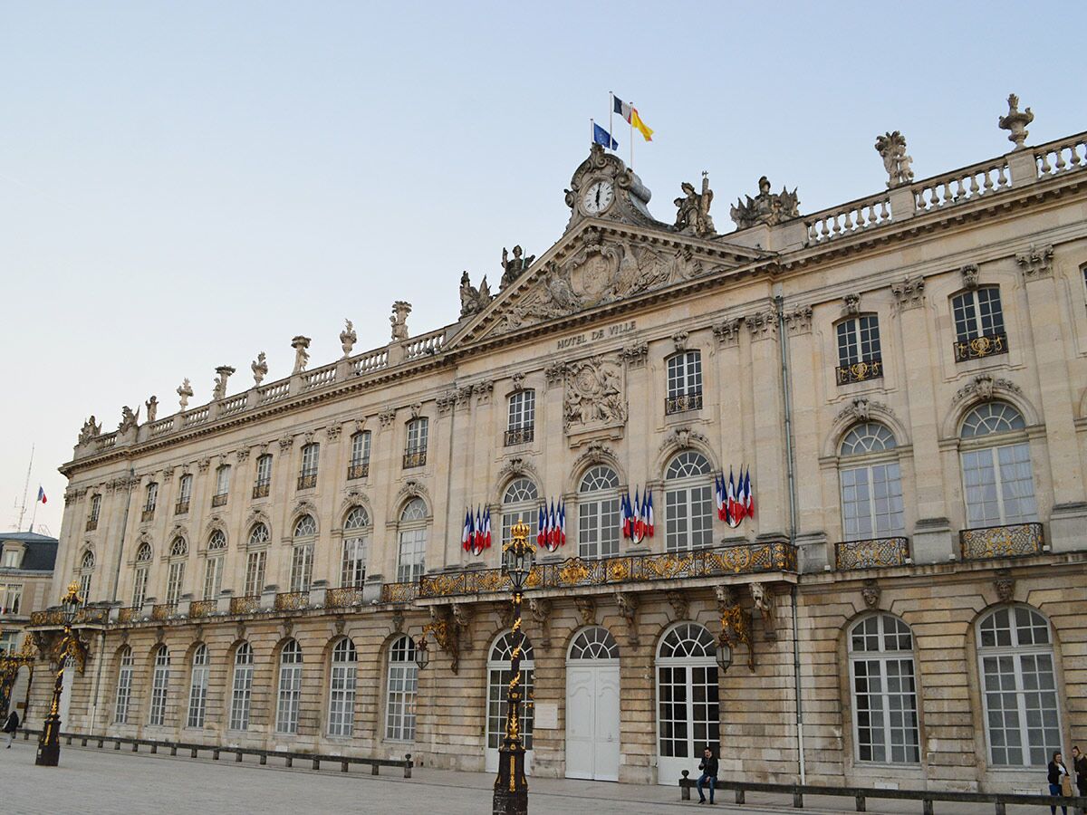 Historisches Hôtel de Ville in Nancy, Frankreich, mit prächtiger Barockfassade, französischen Flaggen und Uhr unter blauem Himmel.