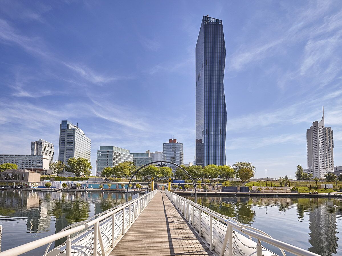 Blick auf die moderne Skyline der Wiener Donaucity mit dem DC Tower 1 und Holzsteg am Wasser unter blauem Himmel.