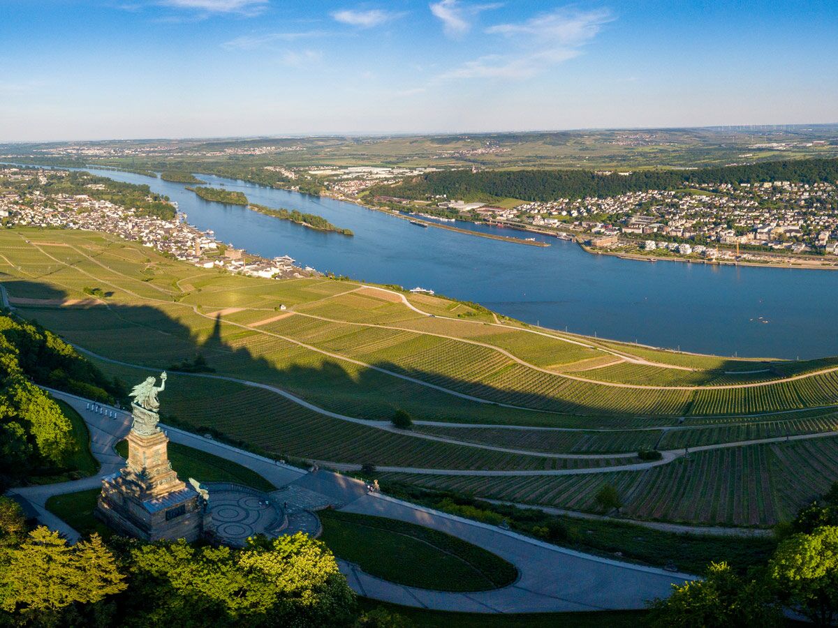 Luftaufnahme des Niederwalddenkmals Germania mit langem Schatten über terrassierten Weinbergen, dem Rhein und Städten.