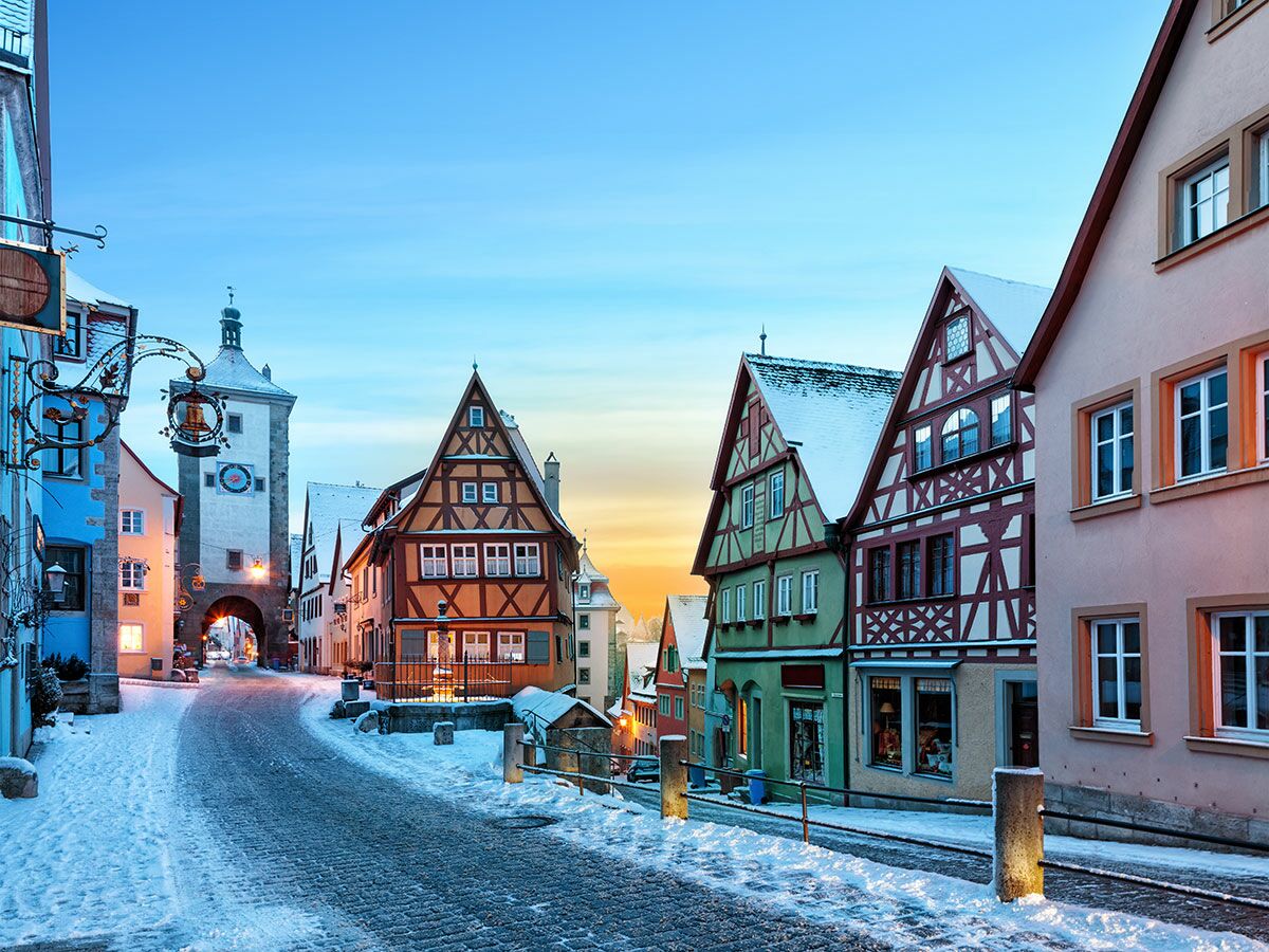 Verschneite Straße in Rothenburg ob der Tauber mit historischen Fachwerkhäusern, Stadttor und Glockenturm bei Dämmerung.