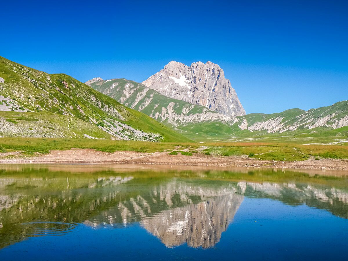 Majestätische Berge mit grünen Hängen spiegeln sich klar in einem ruhigen Bergsee unter blauem Himmel.
