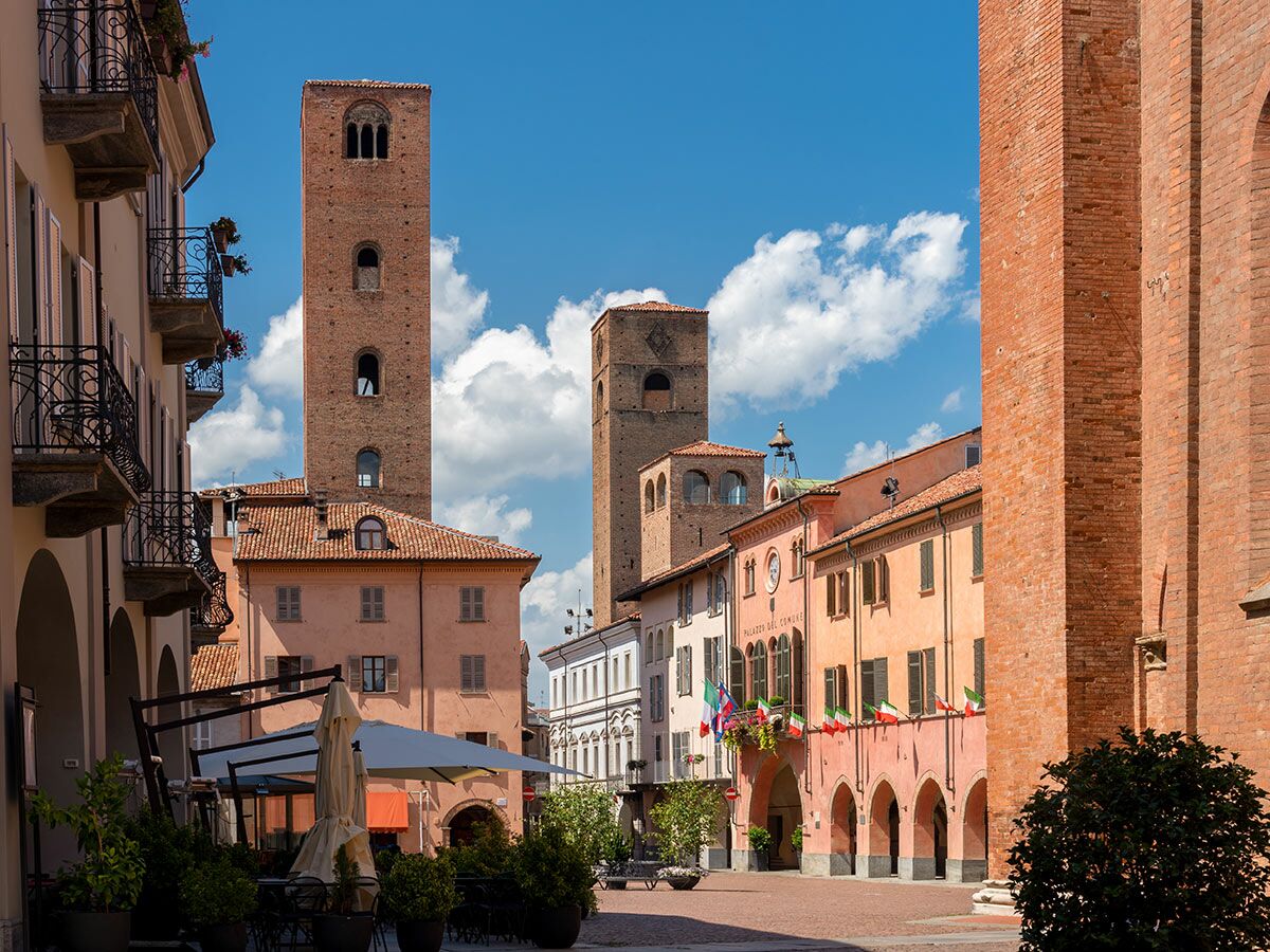 Historischer italienischer Stadtplatz mit hohen Backsteintürmen, traditionellen Gebäuden, Café und italienischen Flaggen unter blauem Himmel.
