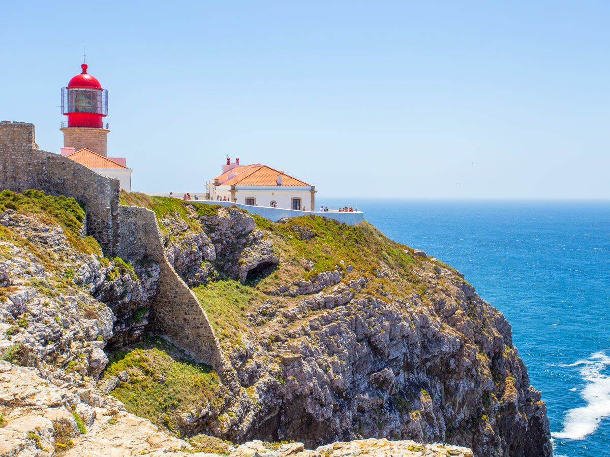 Roter Leuchtturm auf felsiger Steilküste mit historischen Mauern und Gebäuden, Blick auf blaues Meer unter klarem Himmel.