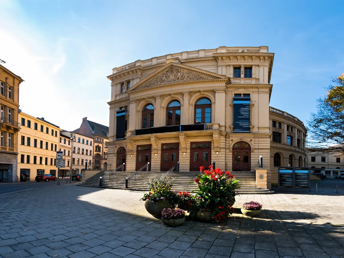 Großes, historisches Theatergebäude mit Steinarchitektur, Treppen, roten Blumen und blauem Himmel an einem sonnigen Tag in der Stadt.