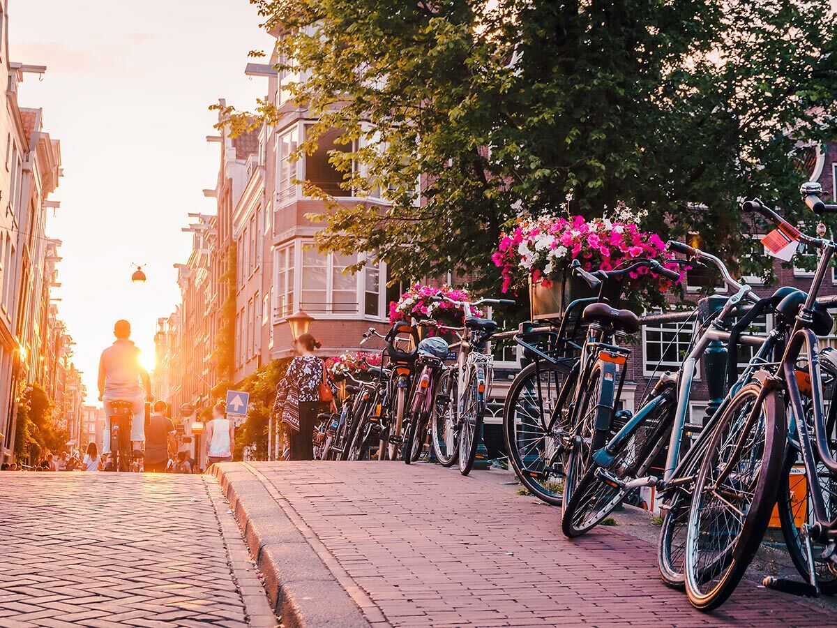 Sonnenuntergang in Amsterdam mit geparkten Fahrrädern an einer Brücke, Kopfsteinpflaster und Menschen auf der Straße.