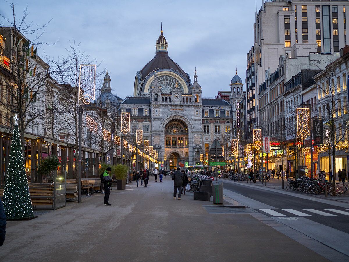 Abendaufnahme des Antwerpener Hauptbahnhofs mit festlicher Weihnachtsbeleuchtung auf der De Keyserlei und Passanten.