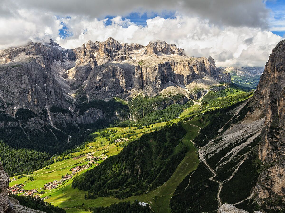 Panoramablick auf die majestätischen Dolomiten mit einem grünen Tal, einem idyllischen Dorf und gewundenen Wegen.
