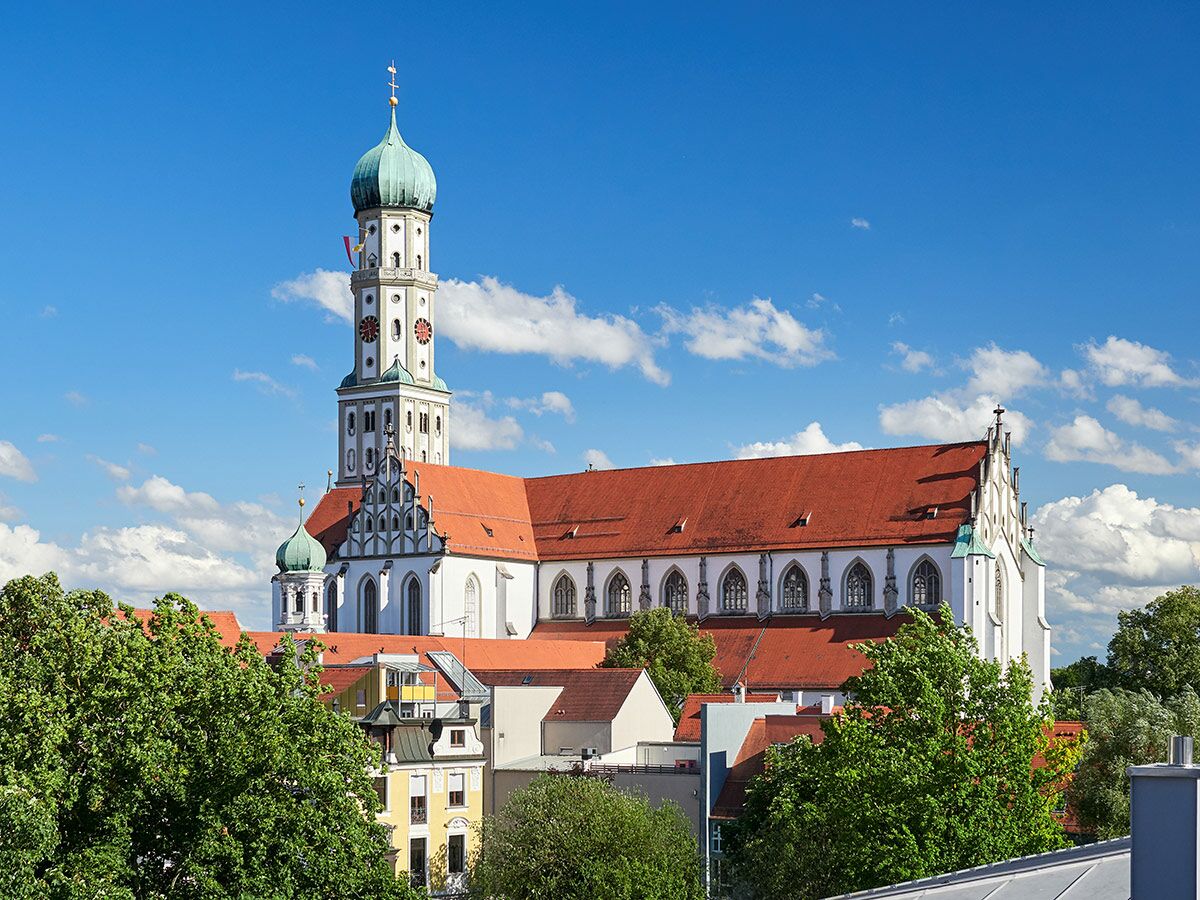 Augsburger Basilika St. Ulrich und Afra mit Zwiebelturm und rotem Dach unter blauem Himmel, umgeben von Bäumen und Gebäuden.