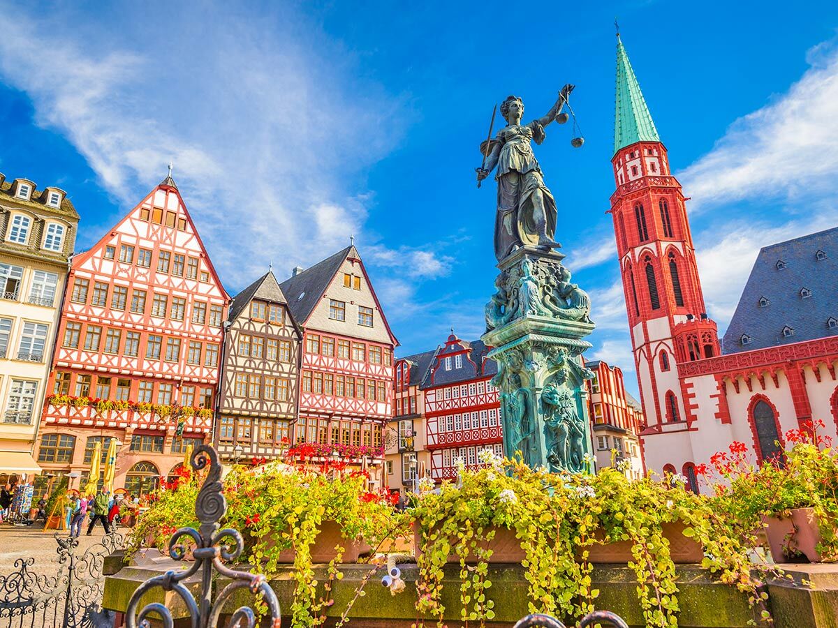 Blick auf Frankfurts Römerberg mit Justitia-Statue, Fachwerkhäusern und Nikolaikirche unter blauem Himmel.