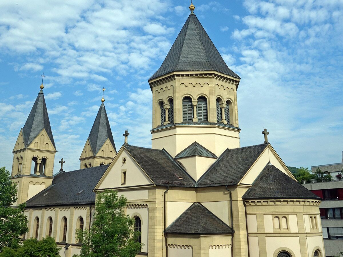 Ansicht einer historischen Kirche mit mehreren Türmen, spitzen Dächern, Kreuzen und hellem Mauerwerk unter blauem Himmel mit Wolken.