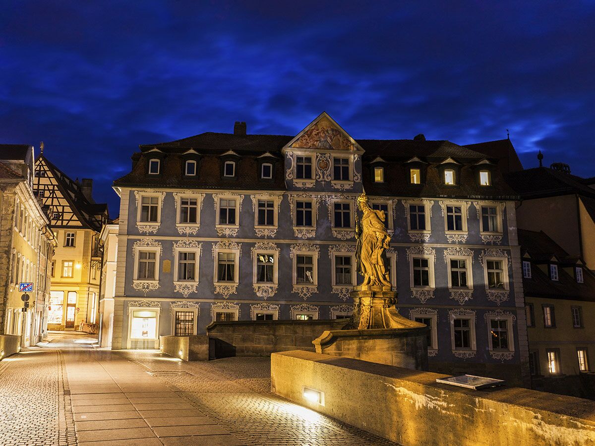 Historische Altstadt von Bamberg bei Nacht mit blauer Fassade, goldener Statue auf Brücke und beleuchteter Gasse.