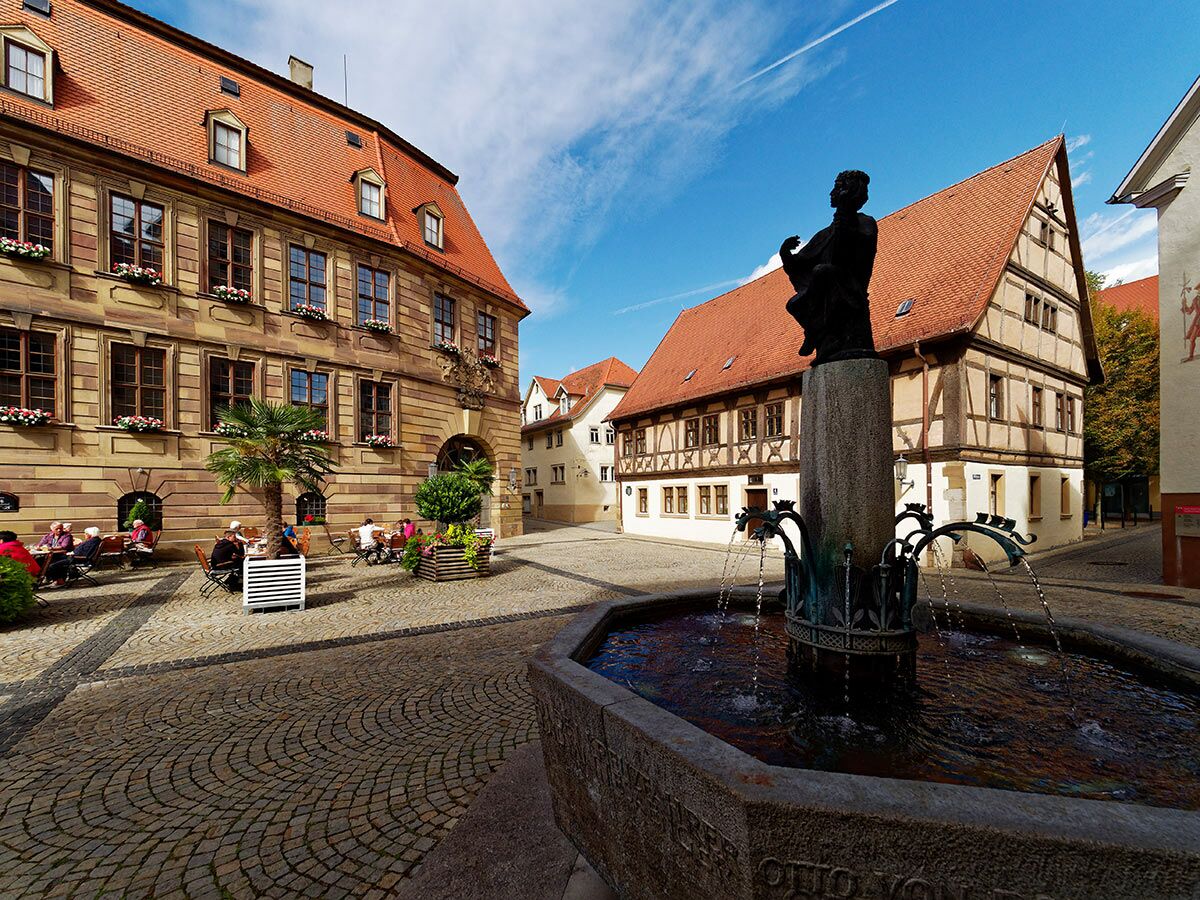 Blick auf einen historischen deutschen Marktplatz mit Steingebäuden, Fachwerkhaus, Springbrunnen mit Statue und Außencafé.