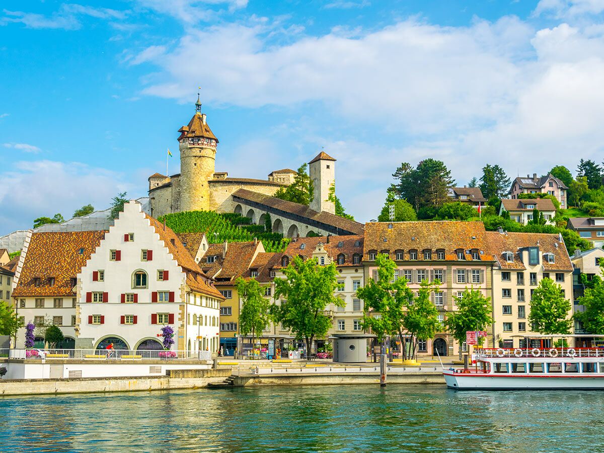 Blick auf eine historische Schweizer Stadt am Fluss mit einem Schloss auf einem grünen Hügel, traditionellen Gebäuden und einem Passagierboot.