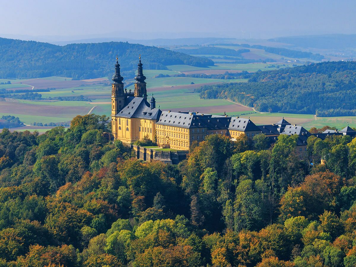 Kloster Banz, ein gelbes Barockschloss, thront auf einem herbstlichen Hügel mit Wald und blickt auf weite Landschaft.