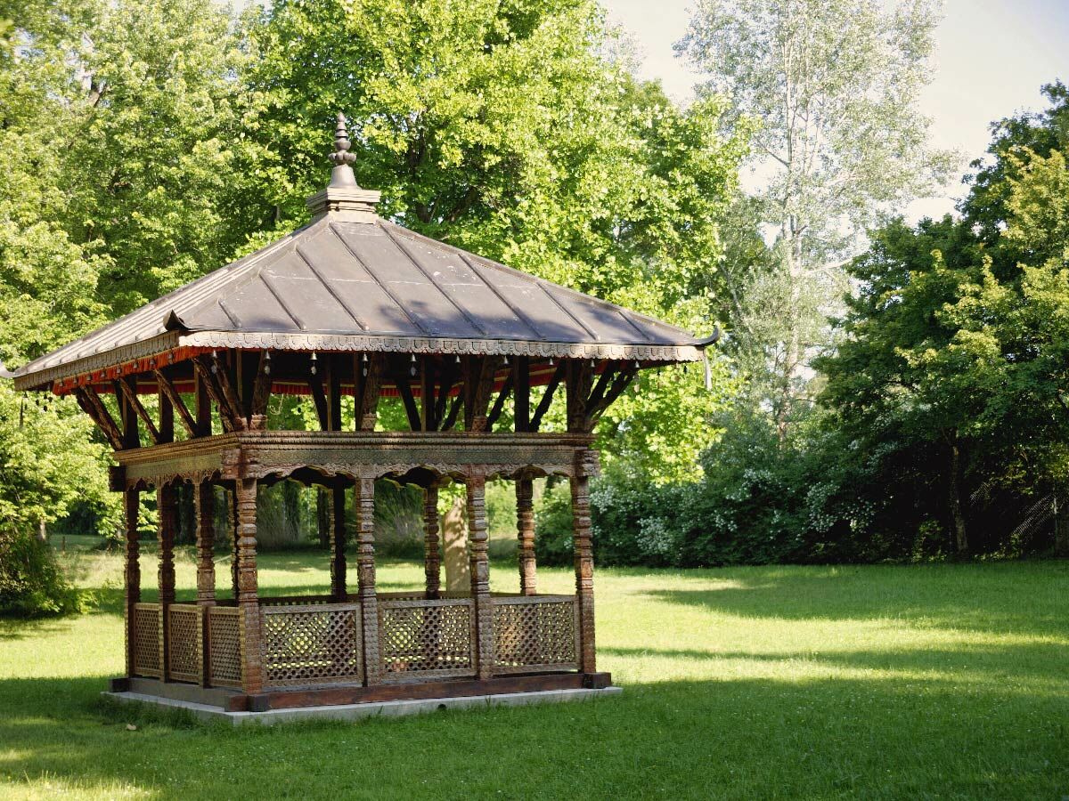 Ein kunstvoll geschnitzter Holzpavillon in einem sonnigen Park, umgeben von dichtem Grün und einem weitläufigen Rasen.
