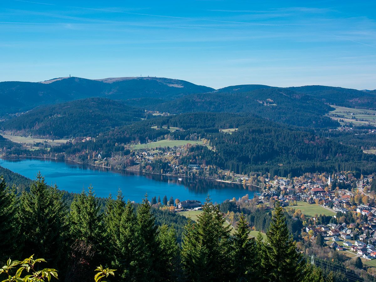 Panoramablick auf den Titisee und Titisee-Neustadt im Schwarzwald, umgeben von Herbstwäldern und Bergen unter blauem Himmel.
