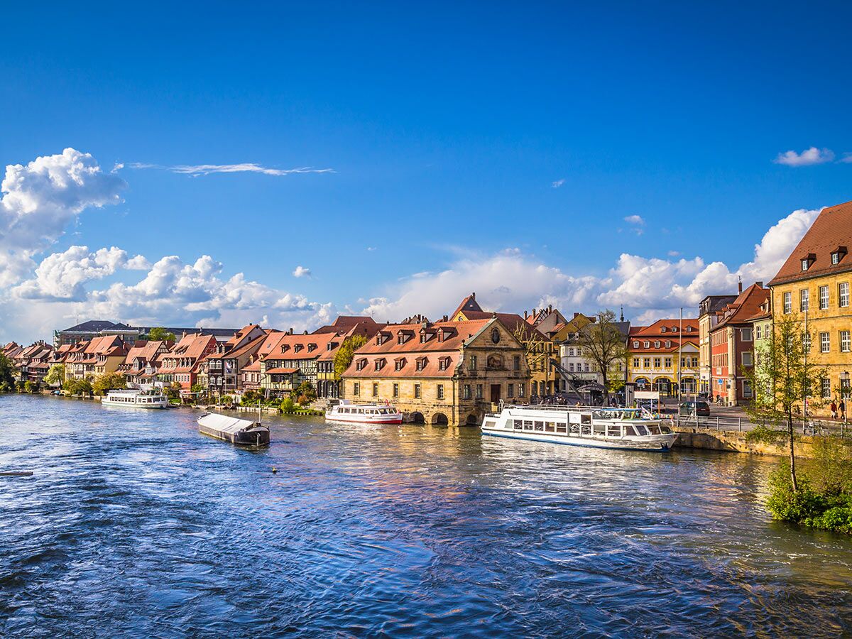 Historische Altstadt von Bamberg am Fluss Regnitz mit bunten Häusern und Touristenbooten unter blauem Himmel.