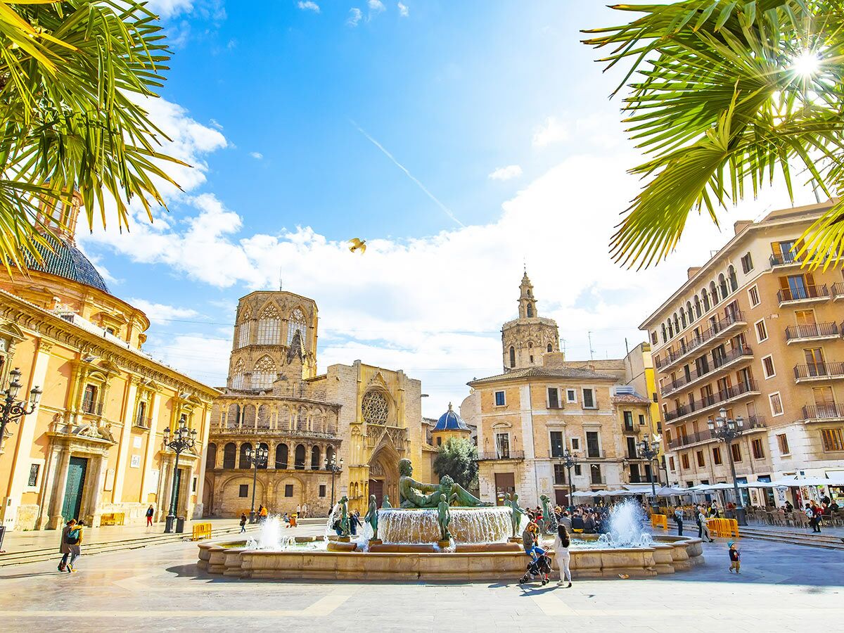 Plaza de la Virgen in Valencia mit Kathedrale, Turia Brunnen, Palmen und Menschen an einem sonnigen Tag.