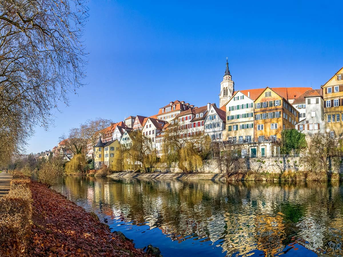 Malerische Neckarfront in Tübingen mit bunten historischen Häusern, Flussreflexionen und Herbstlaub am Ufer unter blauem Himmel.