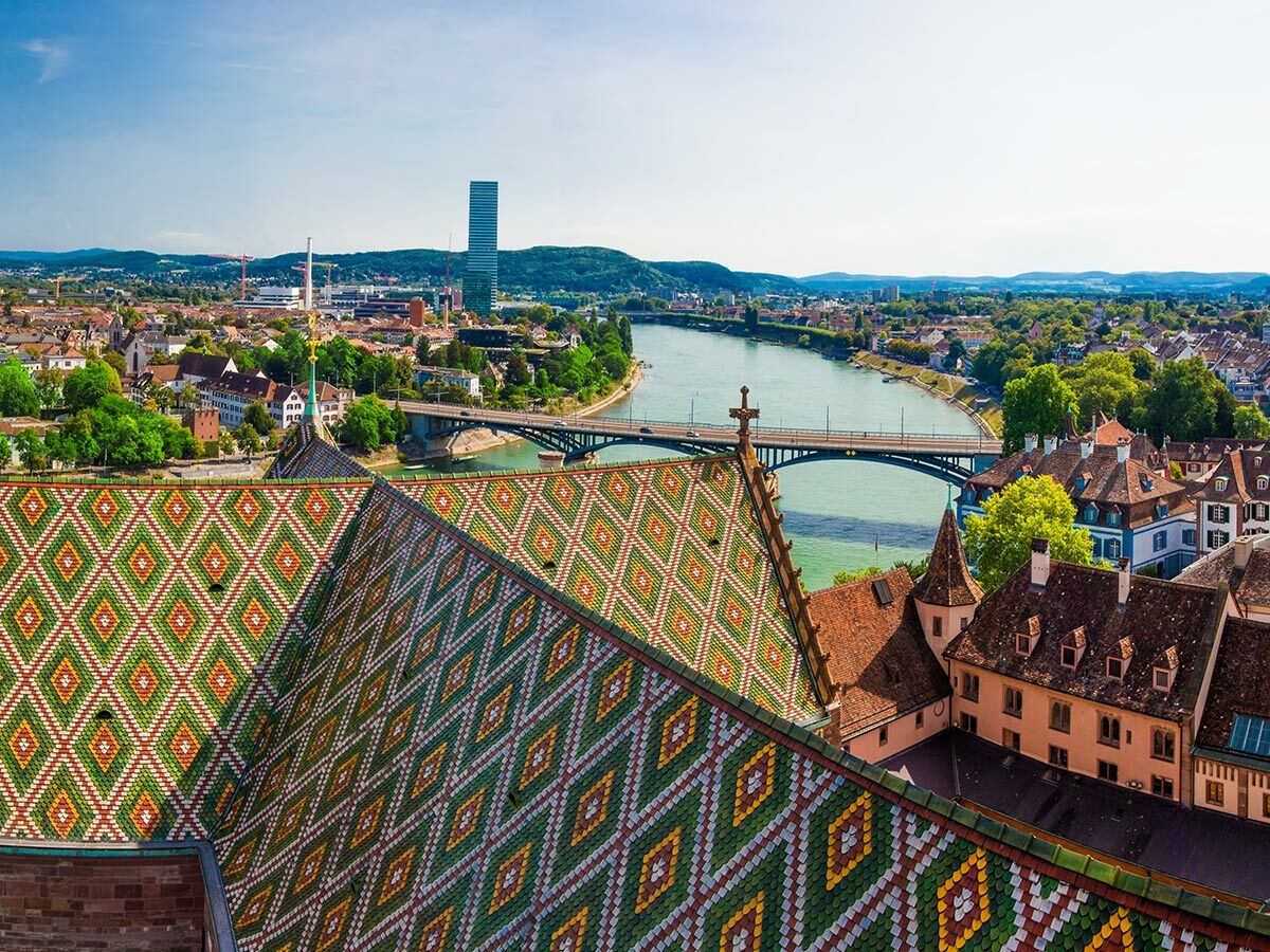 Luftaufnahme Basels mit farbigem Münsterdach, Rhein, Brücken, historischen Gebäuden und Roche Tower bei Sonnenschein.