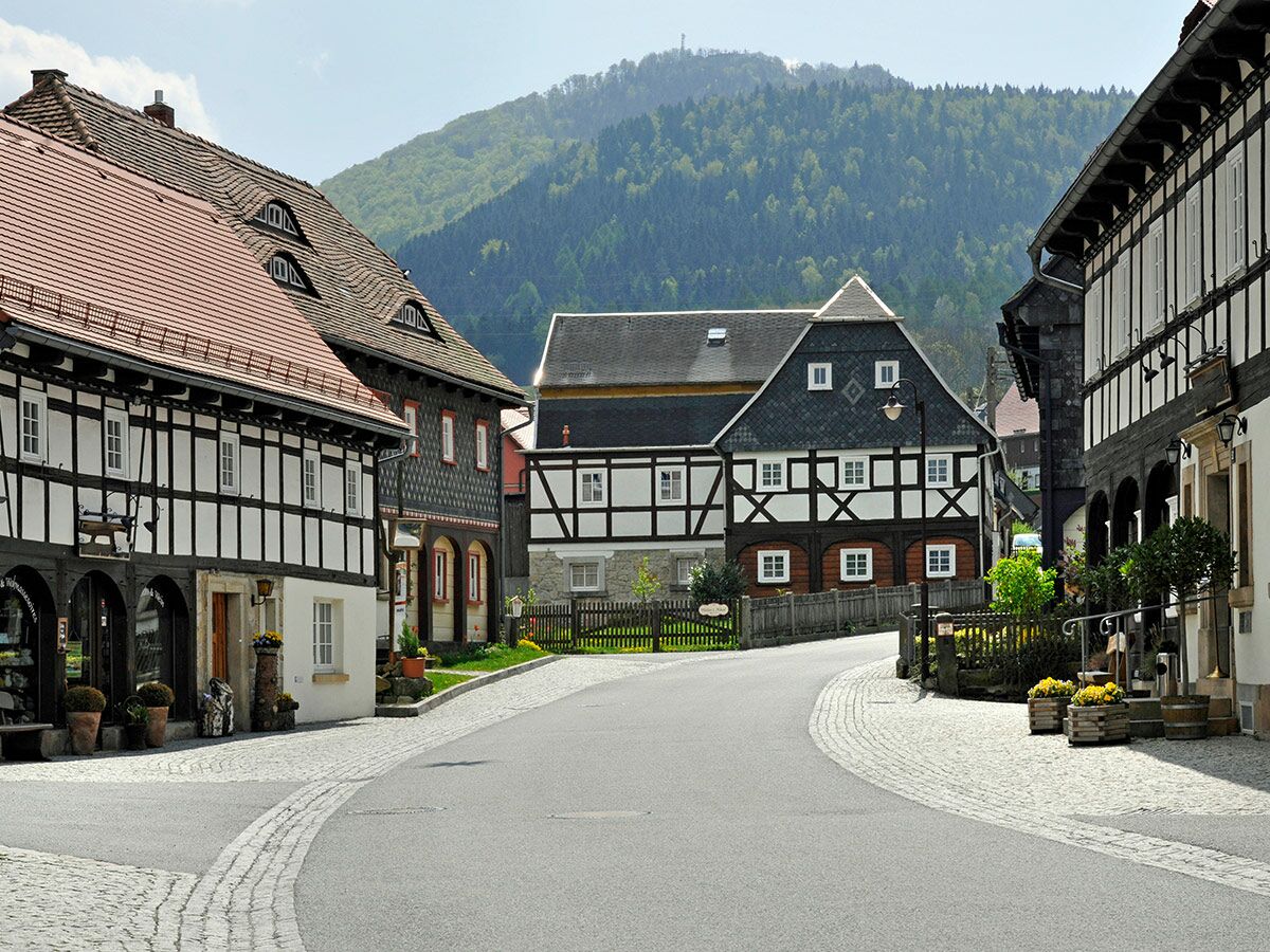 Idyllische Dorfstraße in Deutschland mit traditionellen Fachwerkhäusern und bewaldeten Bergen im Hintergrund.
