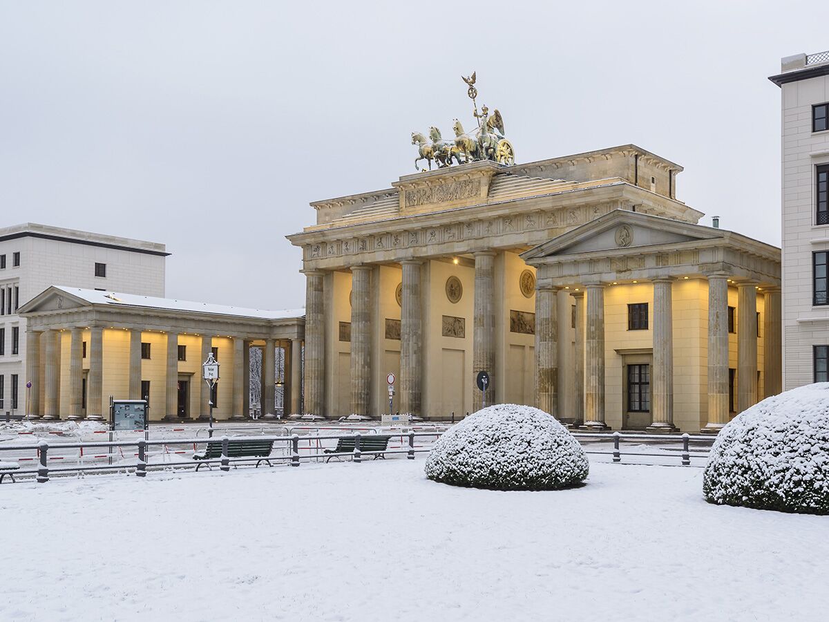 Das Brandenburger Tor in Berlin im Winter, mit Schnee bedeckt und beleuchteten Gebäuden, umgeben von schneebedeckten Büschen und Bänken.