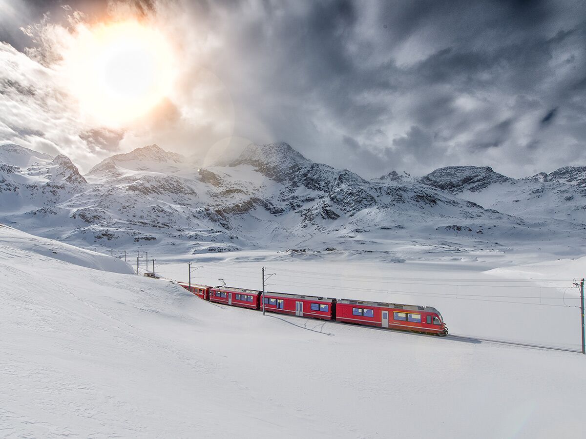 Ein roter Zug fährt durch eine weite, schneebedeckte Berglandschaft. Heller Sonnenschein durchbricht die dramatischen Wolken.