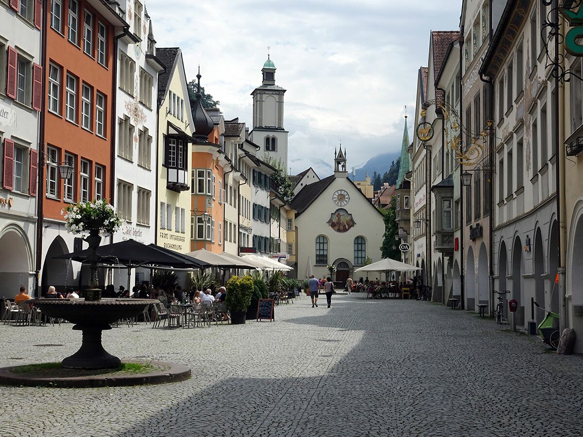 Ein sonniger, kopfsteingepflasterter europäischer Stadtplatz mit bunten historischen Gebäuden, Brunnen und Außencafés.