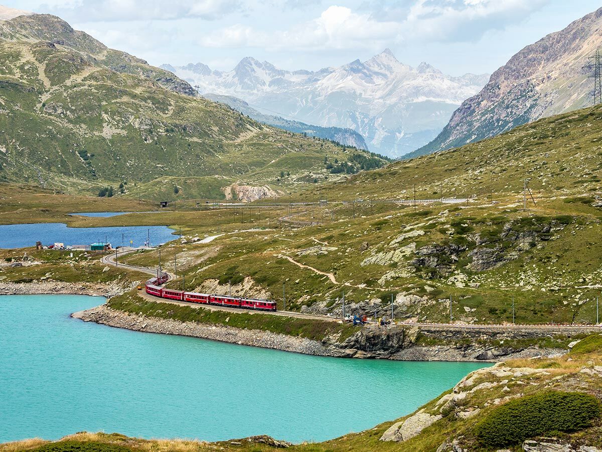 Roter Zug fährt an türkisfarbenem Bergsee entlang, umgeben von grünen Almwiesen und schroffen Alpengipfeln unter bewölktem Himmel.