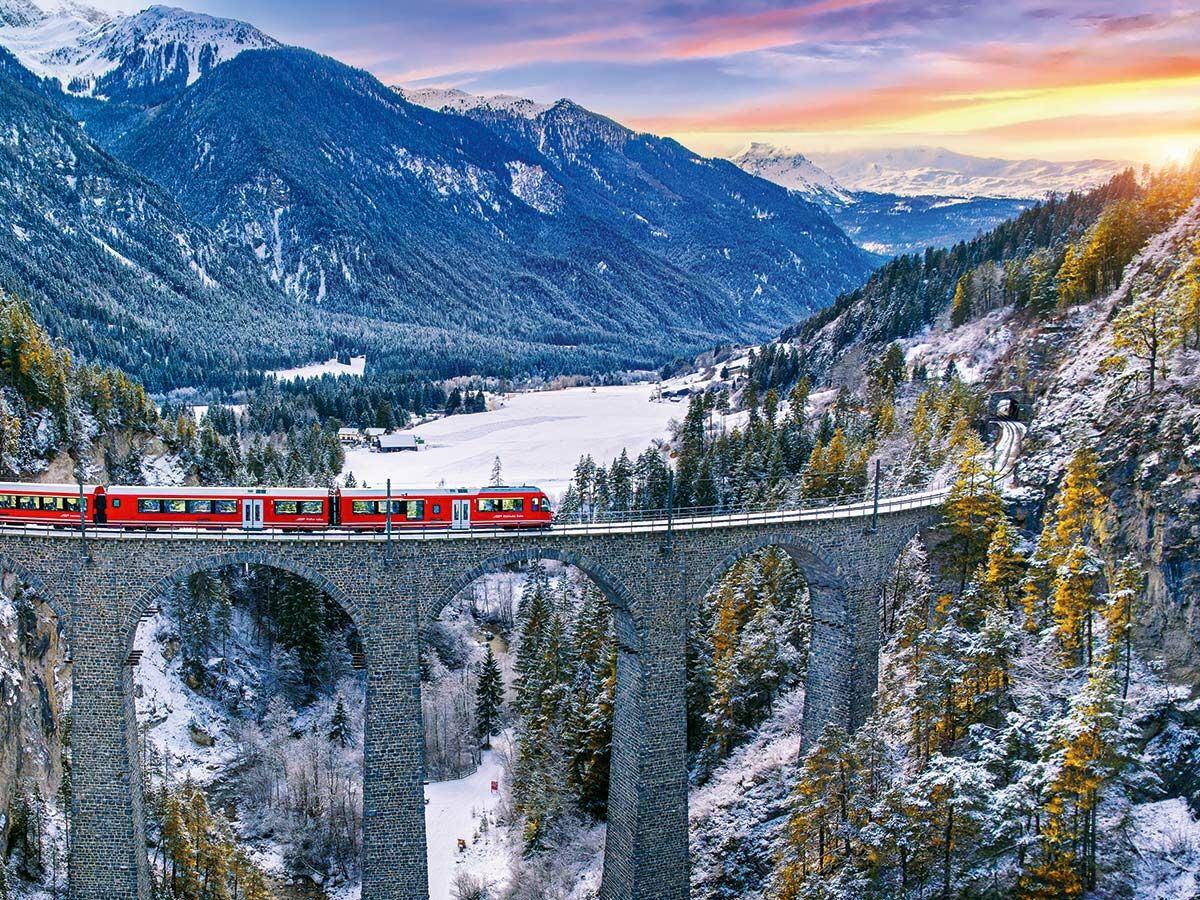 Roter Zug über Landwasser Viadukt in verschneiter Schweizer Alpenlandschaft mit Wald, gefrorenem See und Sonnenuntergang.