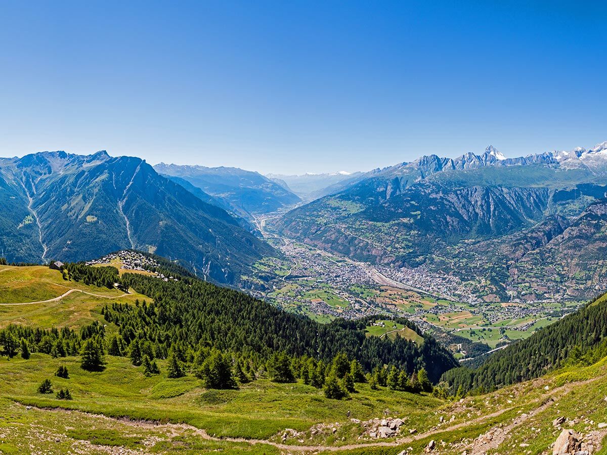 Weitläufiges Panorama der Schweizer Alpen mit einem grünen Tal, einer Stadt, und einem Bergdorf auf bewaldeter Anhöhe unter blauem Himmel.